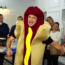 A man in a hot dog costume gestures animatedly while standing in front of a group of people in an office setting, some of whom appear amused or bemused.