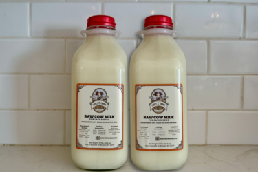 Two clear bottles with red caps, filled with raw cow milk, sit side by side on a countertop against a white tile backsplash. Each bottle is labeled “RAW COW MILK FOR CATS & DOGS.”.