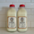 Two clear bottles with red caps, filled with raw cow milk, sit side by side on a countertop against a white tile backsplash. Each bottle is labeled “RAW COW MILK FOR CATS & DOGS.”.