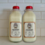 Two clear bottles with red caps, filled with raw cow milk, sit side by side on a countertop against a white tile backsplash. Each bottle is labeled “RAW COW MILK FOR CATS & DOGS.”.