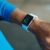 A person wearing a blue long-sleeve shirt checks a white smartwatch on their wrist, displaying a colorful screen with time and fitness activity data.