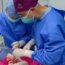Two surgeons in blue scrubs and masks perform a surgical procedure, focusing intently on the operation site, with medical instruments and equipment visible on the table.