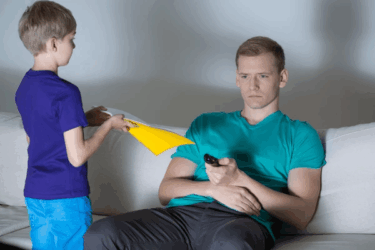 A boy hands a stack of papers to a serious-looking man sitting on a couch, who holds a TV remote and looks away from the boy.