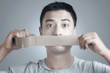 A man holds a strip of duct tape over his mouth, appearing to silence himself. His eyes are wide open, and he wears a light gray shirt against a plain background.
