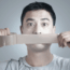 A man holds a strip of duct tape over his mouth, appearing to silence himself. His eyes are wide open, and he wears a light gray shirt against a plain background.