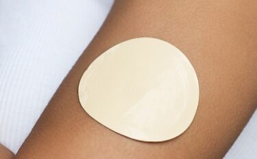 A close-up of a person's upper arm wearing a white sleeveless shirt, with a large beige adhesive patch applied to their skin.