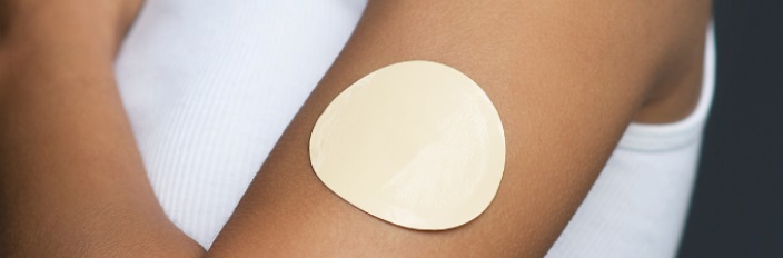 A close-up of a person's upper arm wearing a white sleeveless shirt, with a large beige adhesive patch applied to their skin.