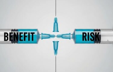 Two syringes filled with blue liquid face each other, labeled "BENEFIT" on the left and "RISK" on the right, with their needles nearly touching in the center.