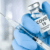 A gloved hand holds a vial labeled "COVID-19 Coronavirus Vaccine" next to a filled syringe, ready for injection. The background is blurred and light-colored.