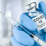 A gloved hand holds a vial labeled "COVID-19 Coronavirus Vaccine" next to a filled syringe, ready for injection. The background is blurred and light-colored.