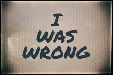 Text on a beige, vertically striped background reads "I WAS WRONG" in bold, black, handwritten-style letters.