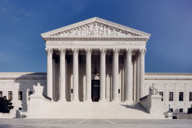 The front view of the United States Supreme Court building in Washington, D.C., featuring tall columns, statues, steps leading to the entrance, and a clear blue sky overhead.