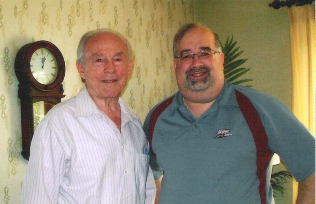 Two men stand indoors smiling at the camera. One wears a light-striped shirt; the other wears glasses and a blue polo shirt. A vintage wall clock and patterned wallpaper are visible in the background.