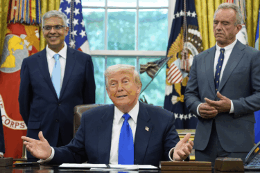 Former President Donald Trump sits at the Oval Office desk, gesturing with open hands, while two men in suits stand behind him smiling. Flags and official emblems are visible in the background.