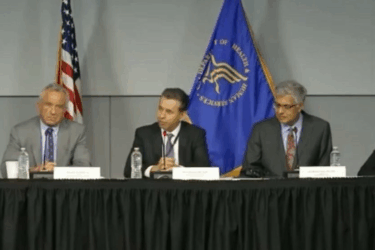 Five men in suits sit behind a table with microphones and water bottles. An American flag and a blue government flag are in the background, indicating a formal panel discussion or press conference.