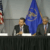 Five men in suits sit behind a table with microphones and water bottles. An American flag and a blue government flag are in the background, indicating a formal panel discussion or press conference.