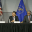Five men in suits sit behind a table with microphones and water bottles. An American flag and a blue government flag are in the background, indicating a formal panel discussion or press conference.