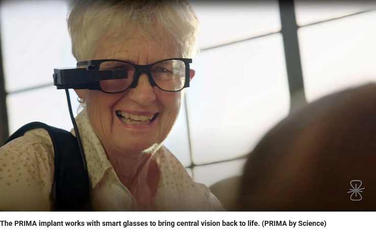 An older woman smiles while wearing smart glasses with an attached device, designed to help restore central vision. Sunlight streams in from a window behind her.