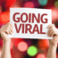 A person holds up a white sign with bold red text reading "GOING VIRAL" against a colorful, blurred background of bokeh lights.