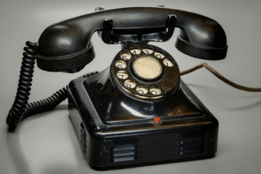 A vintage black rotary dial telephone with a coiled cord sits on a gray surface. The phone has a handset resting on top and a round dial in the center.