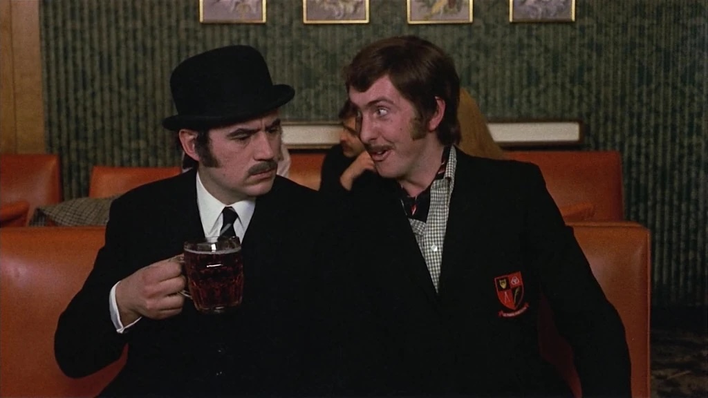 Two men sit on orange chairs in a lounge; one in a black suit and bowler hat holds a glass mug, looking serious, while the other in a blazer with patches smiles and leans in, speaking animatedly.