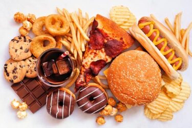 An assortment of fast food and snacks, including a cheeseburger, hot dog, pepperoni pizza slice, french fries, potato chips, chocolate bar, donuts, cookies, popcorn, onion rings, and a glass of soda with ice.