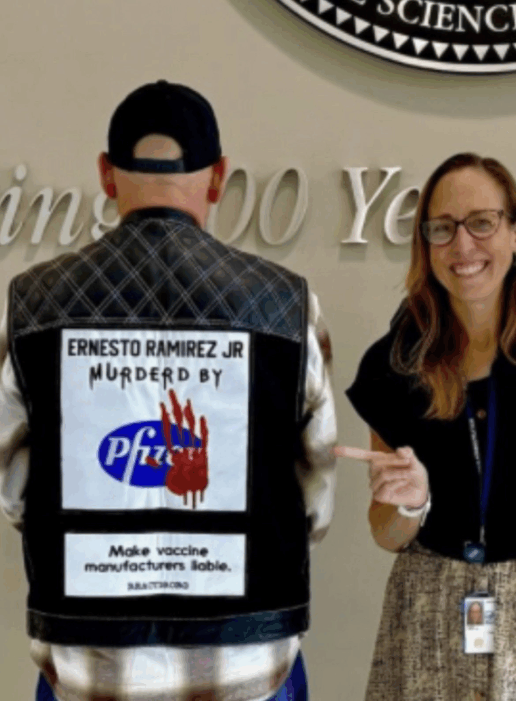 A man wearing a jacket with a sign blaming Pfizer for a death stands next to a smiling woman who is pointing at the sign. The background shows part of a wall sign with large text.