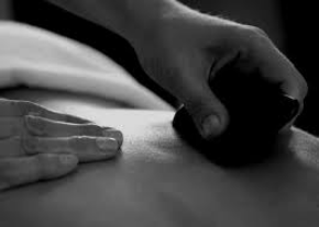A close-up, black and white image shows one hand massaging a person's back while the other hand holds a smooth hot stone, likely during a hot stone massage therapy session.