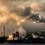 Large factory chimneys releasing thick smoke into the air, with industrial buildings and ships in the foreground under a cloudy sky, indicating heavy air pollution.