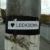 A sticker on a weathered pole reads "I ♥ LOCKDOWN" in black and white. The pole is on a bridge next to an empty street with buildings and trees in the background.