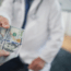 A person in a white lab coat, possibly a doctor, hands several hundred-dollar bills to another person, suggesting a financial exchange in a medical setting.