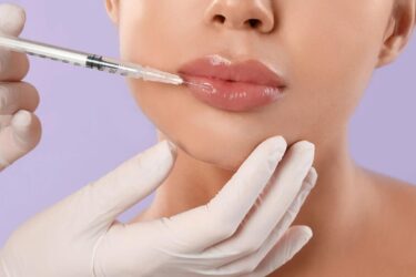 A person with glossy lips receives a lip injection from a gloved hand holding a syringe, suggesting a cosmetic procedure against a light purple background.