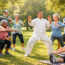 A group of elderly adults exercises together outdoors in a park, following an instructor in white practicing tai chi. Some participants squat or stretch, all smiling and appearing engaged in the activity. Trees and grass are in the background.