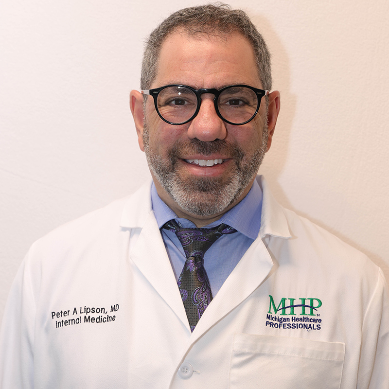 A smiling man with short gray hair, glasses, and a beard wears a white lab coat embroidered with "Peter A Lipson, MD, Internal Medicine" and "MHP Michigan Healthcare Professionals.