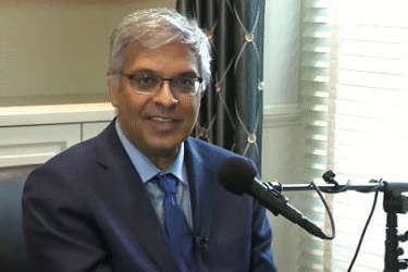 A man in a blue suit and tie sits indoors, smiling at the camera while speaking into a microphone. There is a window with blinds and a curtain in the background.
