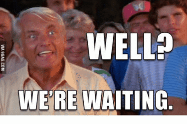 An older man with white hair, wide eyes, and a tense expression stands in front of a crowd. Large bold text reads, "WELL? WE'RE WAITING." The image conveys impatience or eagerness for a response.