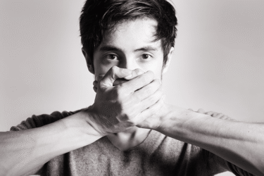 A young man in a grey shirt covers his mouth with both hands, looking directly at the camera. The image is in black and white with a plain background.