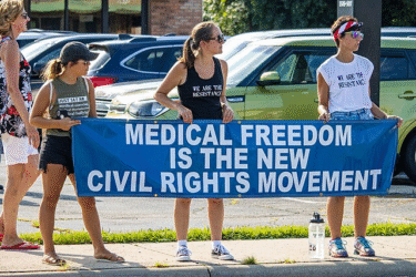 Four people stand on a sidewalk holding a large blue banner that reads, "Medical Freedom Is The New Civil Rights Movement." Two women wear shirts with protest slogans. Cars are parked in the background.