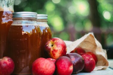 Several red apples spill from a burlap sack next to jars of apple cider on an outdoor table, with a blurred green background.