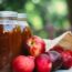 Several red apples spill from a burlap sack next to jars of apple cider on an outdoor table, with a blurred green background.