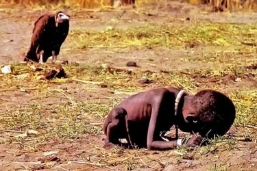 A malnourished child crouches on dry ground with their head down, while a vulture stands in the background, watching from a short distance. The scene is barren and evokes a sense of hardship and desperation.