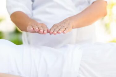 A person holds their hands hovering above another person’s body, suggesting a healing or energy therapy session, both dressed in white with a blurred outdoor background.