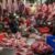 A butcher wearing a red shirt cuts raw meat at a market stall, surrounded by slabs of meat hanging overhead and trays of meat on the counter. The workspace is busy and filled with fresh cuts.