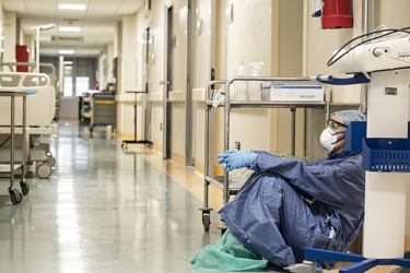 A healthcare worker in full protective gear sits on the floor against a hospital wall, looking tired. Medical equipment and empty beds are visible in the hallway. The scene suggests exhaustion during a busy shift.