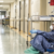 A healthcare worker in full protective gear sits on the floor against a hospital wall, looking tired. Medical equipment and empty beds are visible in the hallway. The scene suggests exhaustion during a busy shift.
