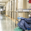 A healthcare worker in full protective gear sits on the floor against a hospital wall, looking tired. Medical equipment and empty beds are visible in the hallway. The scene suggests exhaustion during a busy shift.