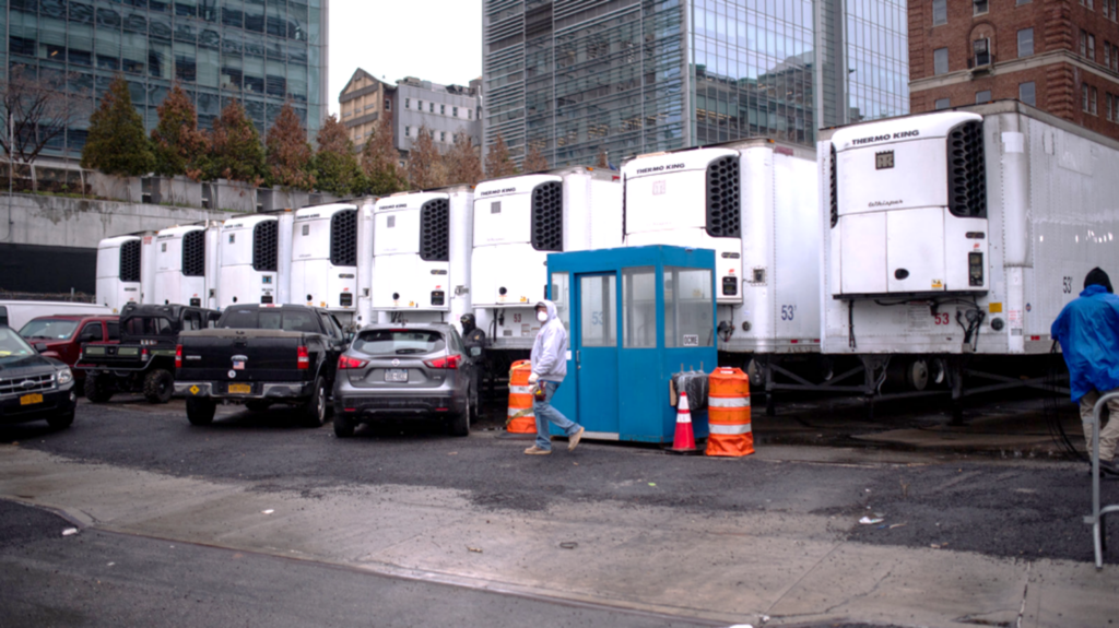 A row of refrigerated trucks is lined up in a city lot next to parked cars. A person in a white coat and mask walks past a small blue guard booth, with tall buildings visible in the background on a cloudy day.