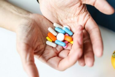 A pair of hands holds a variety of colorful pills and capsules, including white, blue, orange, and pink medications, against a light background.