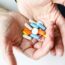A pair of hands holds a variety of colorful pills and capsules, including white, blue, orange, and pink medications, against a light background.