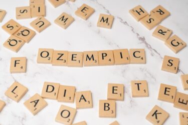 Scrabble tiles spell out the word "OZEMPIC" on a white marble surface, surrounded by scattered tiles with random letters.